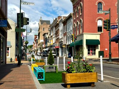 Colorful wooden benches, planter boxes filled with flowers and small shrubs, and patches of artificial turf occupy the converted travel lane on West Beverley Street at New Street, separated from traffic by white flexible delineator posts, with downtown Staunton's historic brick storefronts lining both sides of the street.