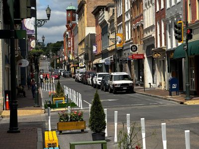 An elevated view looking west down Beverley Street from New Street, showing the full two-block demonstration corridor with colorful benches, flower-filled planter boxes, evergreen shrubs, and artificial turf patches lining the converted travel lane, with historic downtown Staunton buildings on both sides.