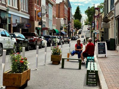 Two people sit on colorful wooden benches in the Beverley Street pedestrian demonstration area, with wooden planter boxes filled with flowers and white flexible delineator posts separating the space from the adjacent travel lane and parked cars, downtown storefronts visible in the background.