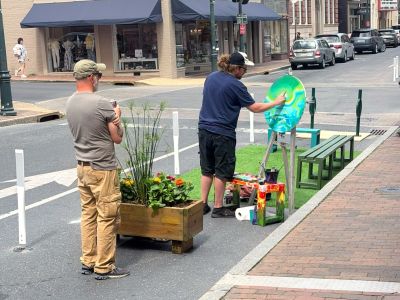An artist paints a colorful circular canvas on an easel in the Beverley Street pedestrian demonstration space, while a bystander watches, with a flower planter, artificial turf, benches, and downtown Staunton storefronts visible behind them.